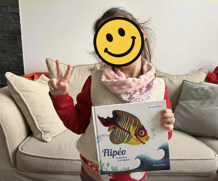 Flipeo le poisson courageux dans les mains d'une heureuse enfant lectrice. Album jeunesse dès 5 à 6 ans et pour tous les amoureux de la nature, des mers et des océans. Une aventure amusante qui parle de nature, d'environnement et d'amitié. Un livre au format carré 21 x 21 cm, 40 pages, couverture rigide. Textes par Hervé Hernu, illustrations et mise en page par Aline Hernu, aux éditions La Petite Fabrique de Livres (par Nathalie Clairet Boitel)