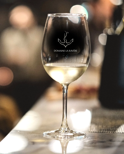 Logo du Domaine La Ravère sur un verre de vin blanc. Identité visuelle conçue pour Paul Brunet - artisan vigneron bio. Vignoble en Drôme Ardèche (Auvergne Rhône Alpes). Design par Aline Hernu - Studio AirNewArt. 2025, tous droits réservés.